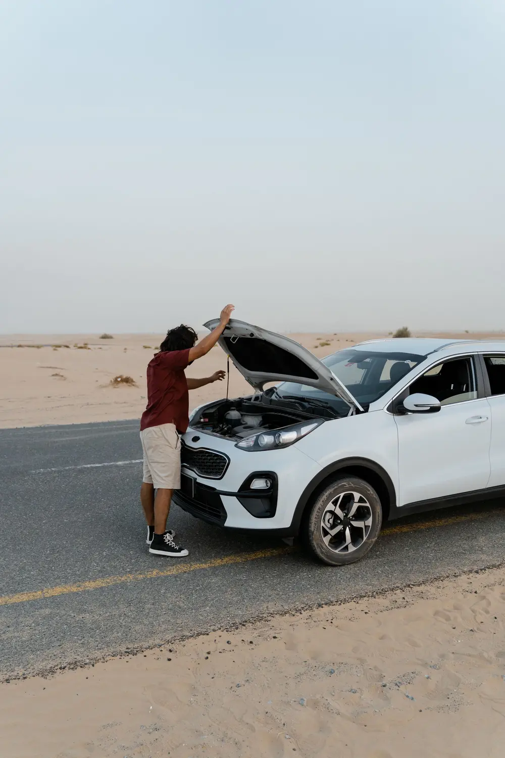 A driver looking under the raised hood of a white SUV stopped on a desert road