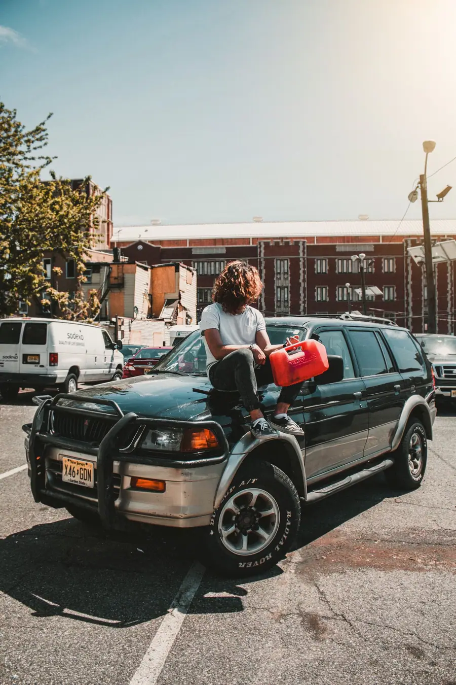 A driver sitting on the hood of an SUV in a parking lot, holding a red gas can