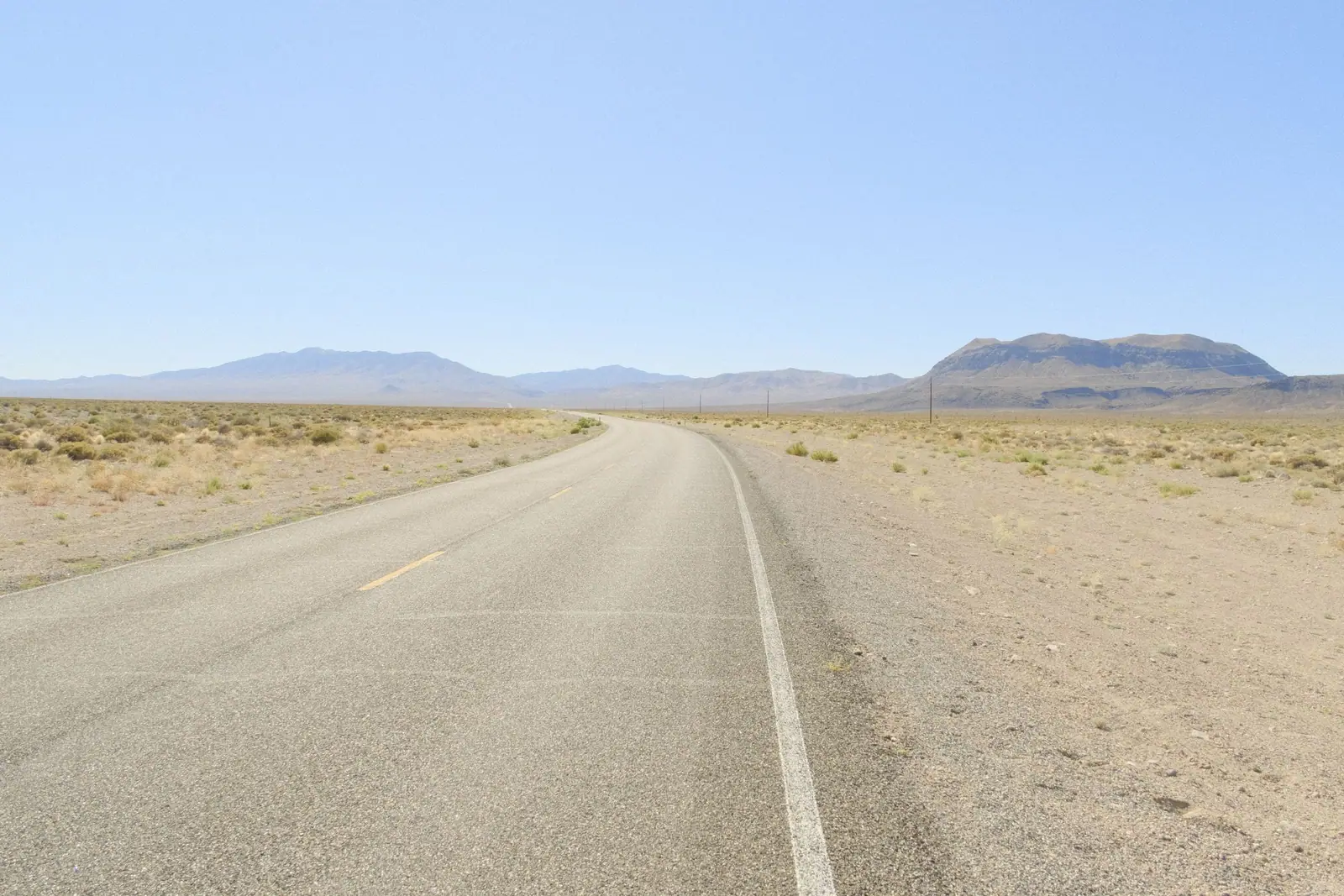 An empty two-lane desert highway in Nevada running toward the mountains under a clear sky
