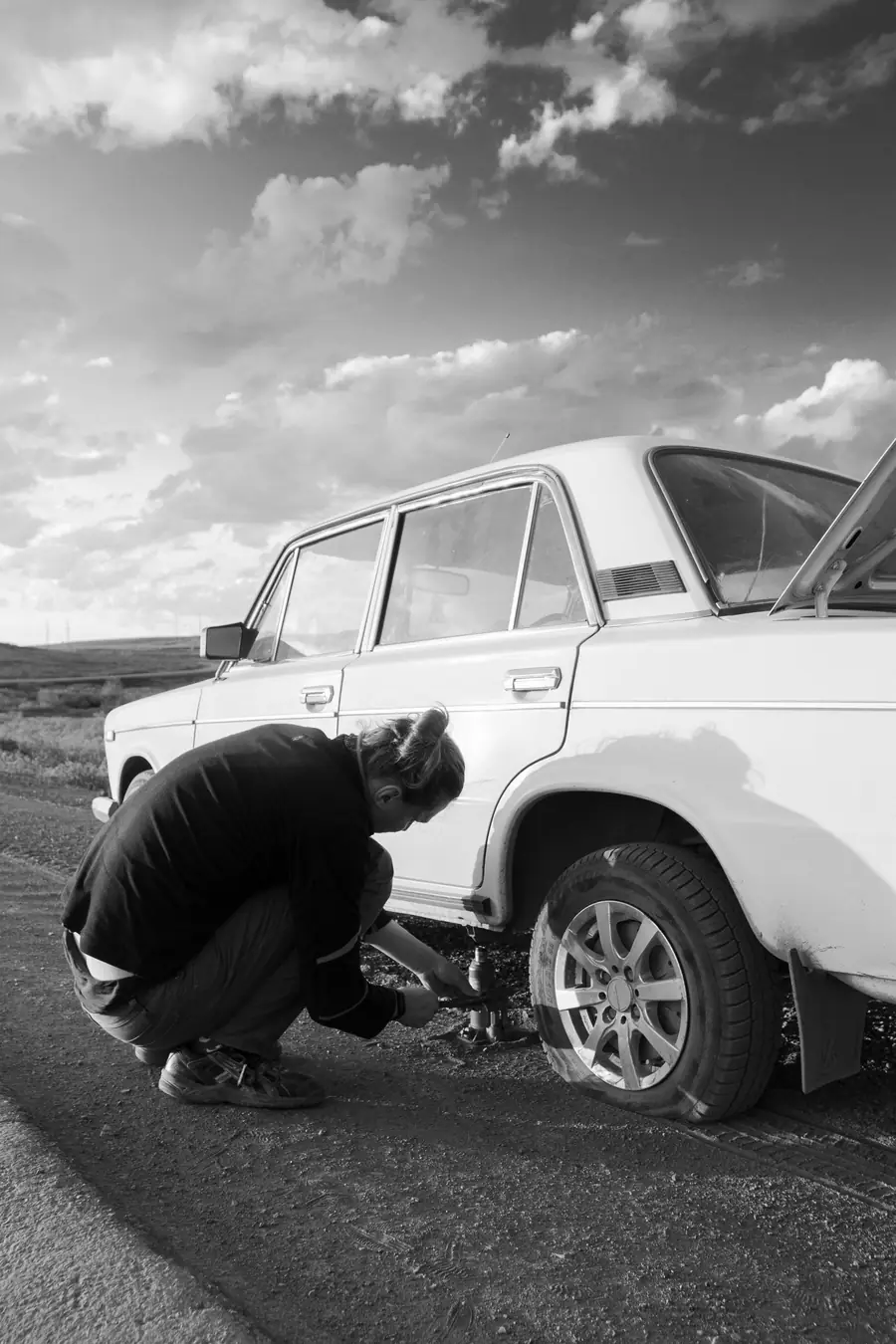 Black-and-white photo of a driver crouched at a car wheel, changing a flat tire at the roadside